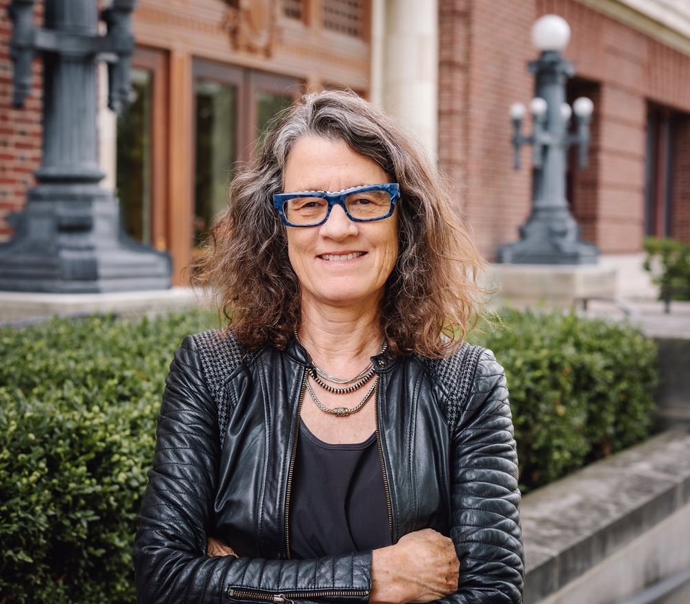 A photo of Dr. Leslie Reagan. She is a white woman standing in front of a row of hedges outside a brick building. She has shoulder-length dark hair and is wearing glasses, a black top, and a black leather jacket. 