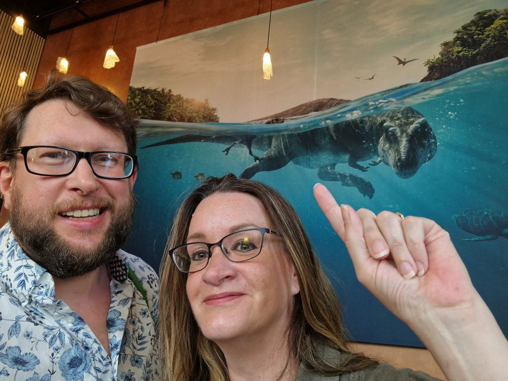 Darren and Toni with giant picture of Prehistoric Planet swimming T. rex behind.