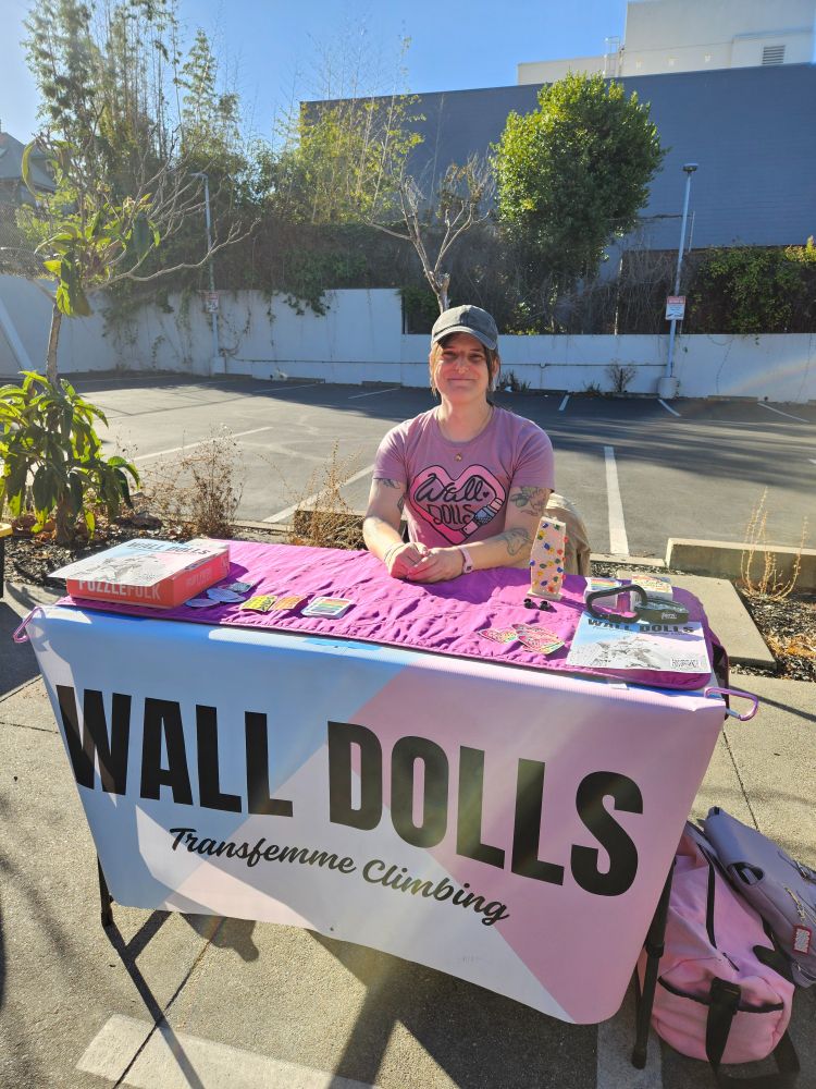 Jamie is sitting in a parking lot at a table. The table has the Wall Dolls Transfemme Climbing banner. She has a Wall Dolls t-shirt on and is smiling.
