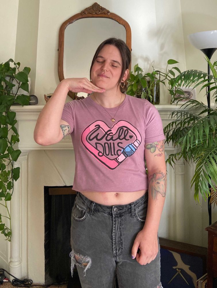 Jamie is wearing a pink crop top with a heart shaped carabiner on it with WALL DOLLS in the center.