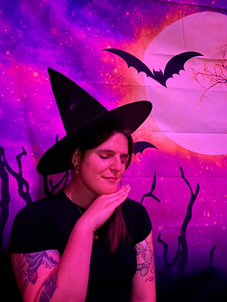 Jamie is wearing a witch's hat in front of a Halloween photo backdrop that features a bat and the moon