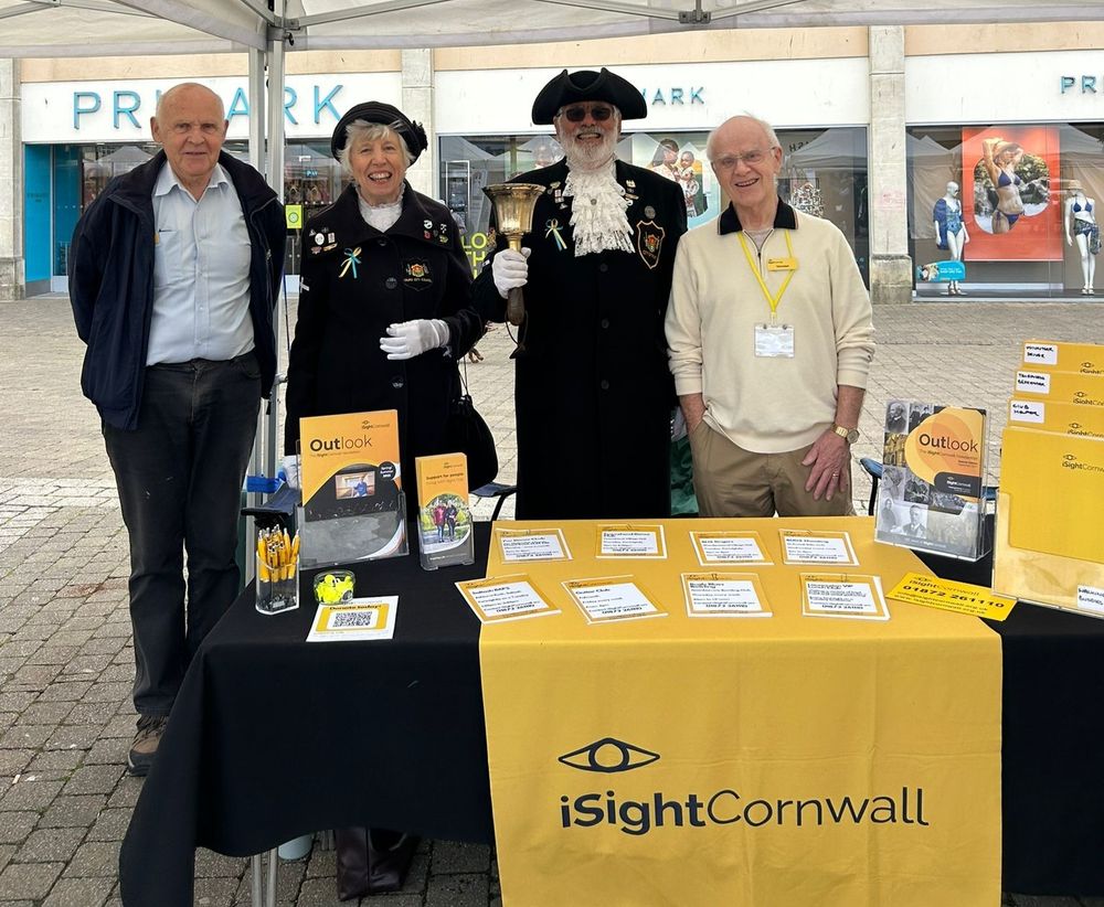 Four people stand behind a table covered in iSightCornwall branding and leaflets. Two are wearing regalia and white gloves, the man holding a bell. Either side of them are two men who are wearing iSightCornwall lanyards and badges.