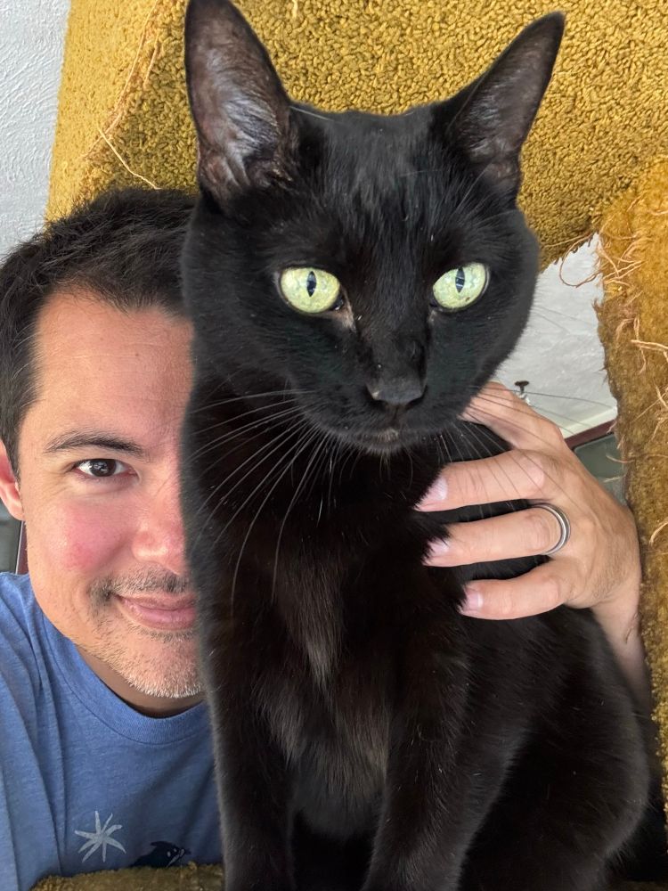 A selfie of a middle aged man posing with a black cat. The cat looks off in the distance but the man looks at the camera. He keeps one hand on her back in case she decides to flee.