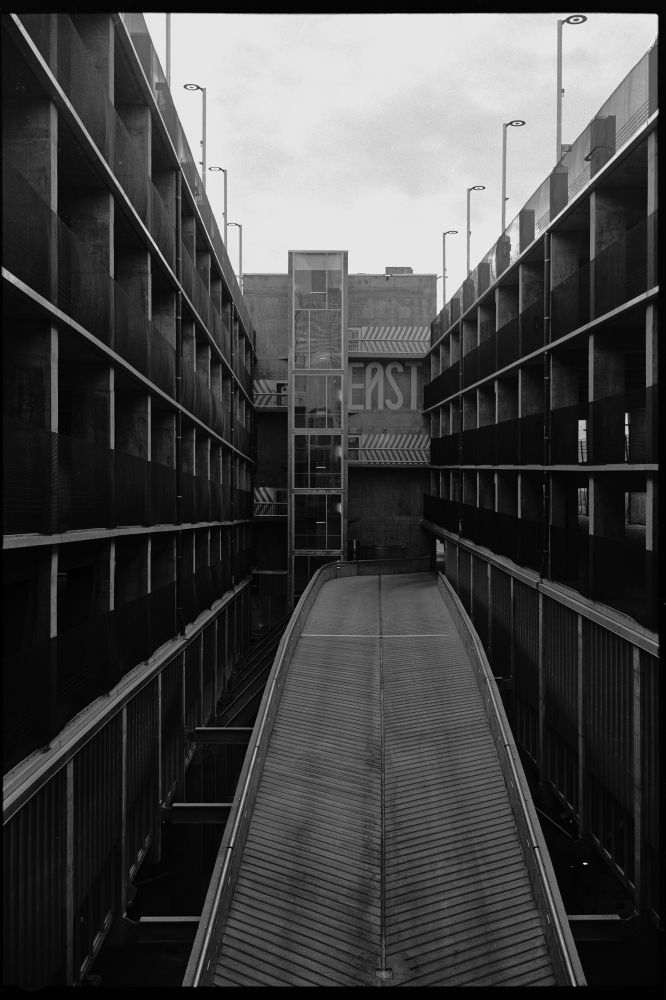 Black and white photograph of a parkade in downtown Calgary, Canada. A concrete ramp rises up from the bottom of the photograph towards a tower and stairwell at the end. EAST is painted in large letters on the far side of the parkade
