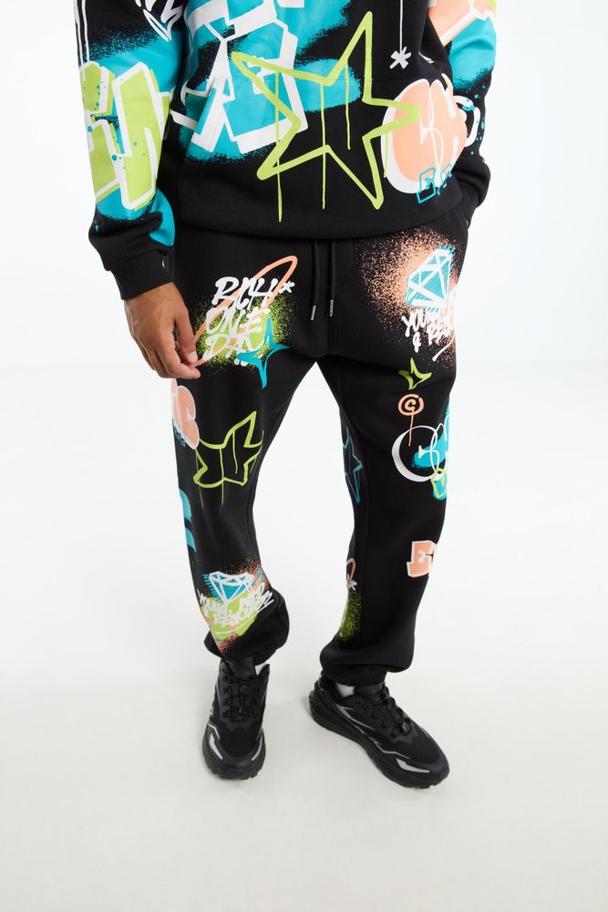 product image of the matching jogging pants. the picture shows the model wearing the set from the bellybutton downwards to the shoes, to showcase the pants. it matches the hoodie with a black base color and a graffiti print all over with the color palette using white as accent color on a cyan blue, lime green and pale orange.