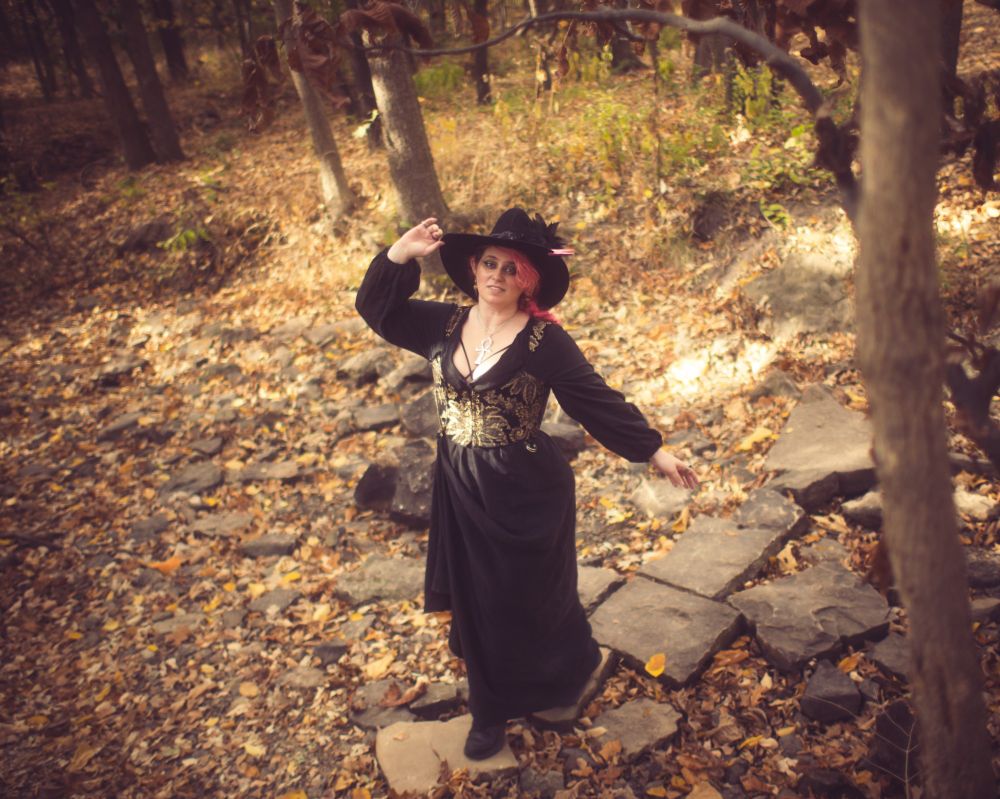a pink-haired woman models a witch hat and handmade black dress with gold and black brocade stays that have POCKETS while enjoying a leaf-littered autumn day in nature