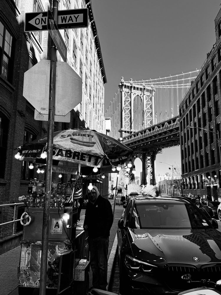 A monochrome photo of a street vendor selling hot dogs in DUMBO. Cars parked along the street with the Manhattan Bridge in the background. 