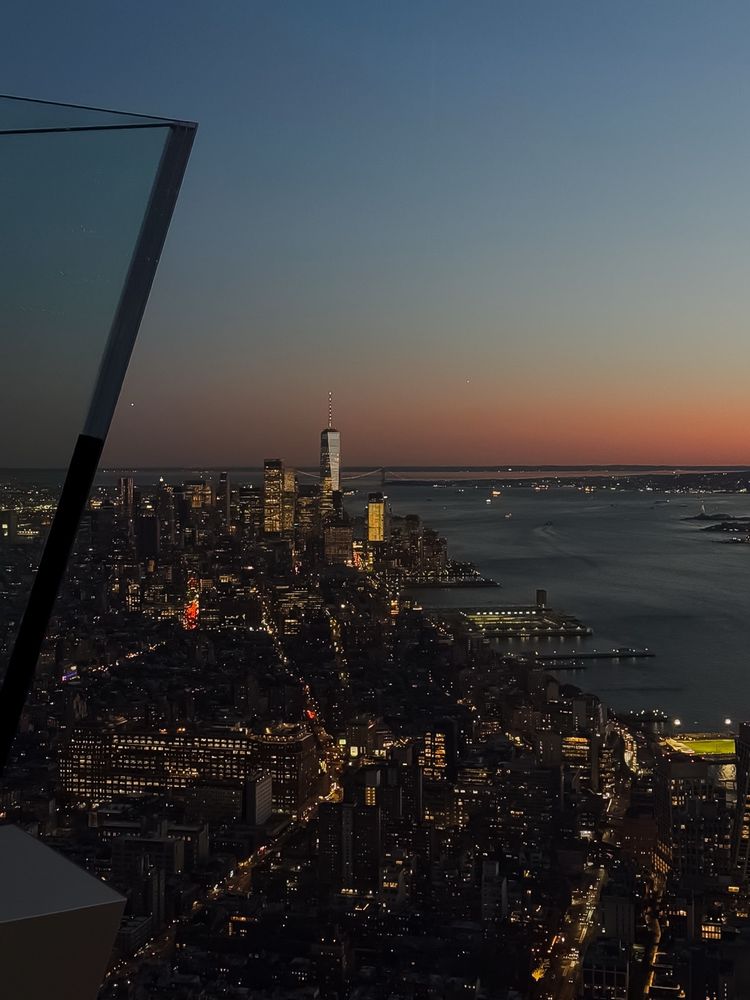 View of downtown Manhattan and the One World Trade Center at dusk, seen from the Edge at Hudson Yards, with city lights glowing beneath.
