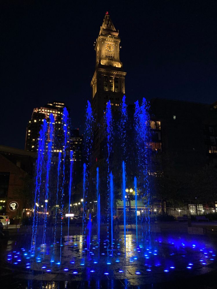 A nighttime scene featuring a brightly lit fountain with blue water jets in the foreground, alongside a historic clock tower illuminated against a dark sky. Urban buildings are partially visible in the background.