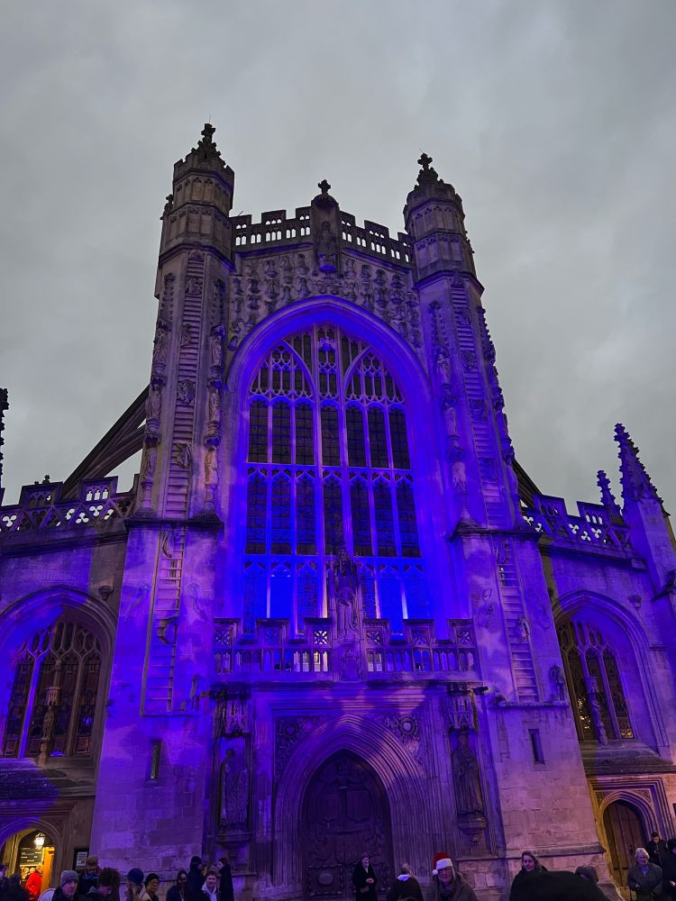 The front of the Bath Abbey illuminated in blue and red star pattern dancing around the light with the night sky in the background. 