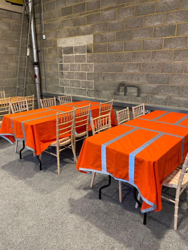 2 tables with completed custom tablecloths and chairs. The walls are exposed grey concrete. The tablecloths are orange with silver reflective trim down each side and trim on the hem. 