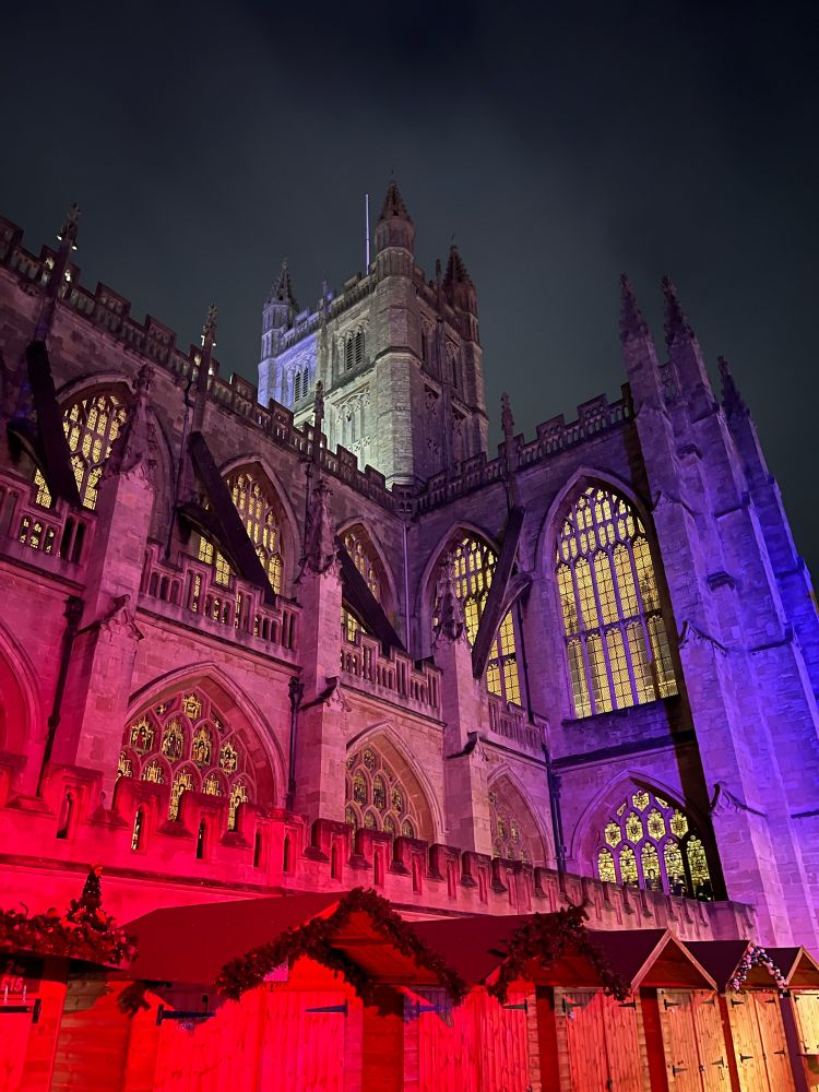 The side of the Bath Abbey beautifully lit in a mixture of red and purple with the dark night sky in the background. At the bottom of the photo are rows of wooden stalls closed for the night during the Christmas market. 