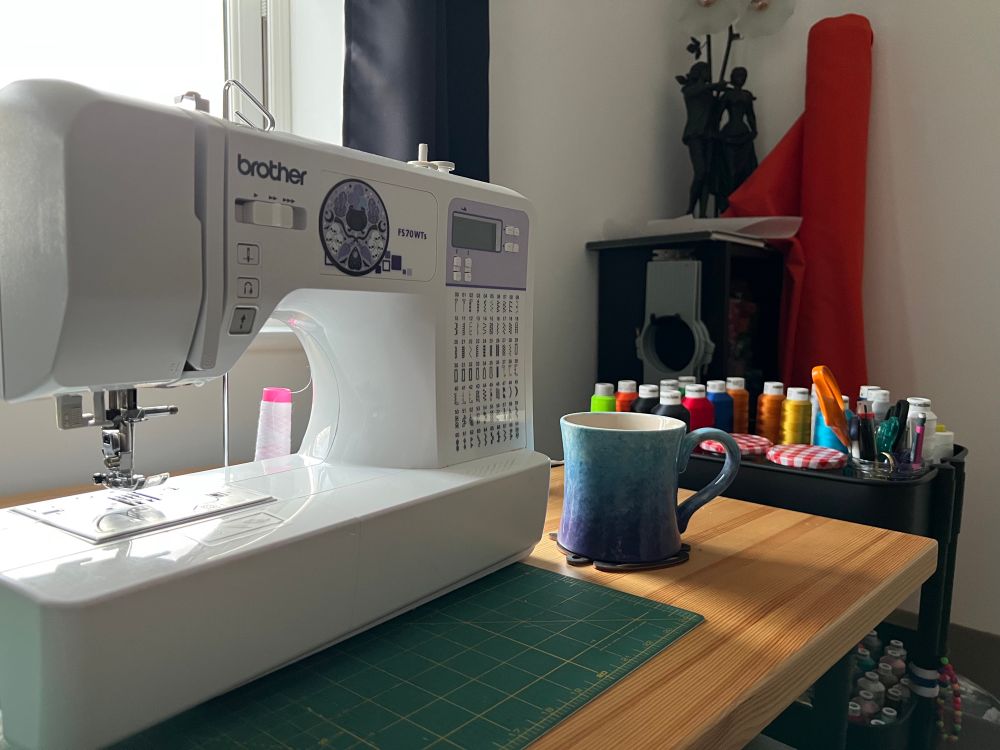 White sewing machine and blue ombré coffee mug on a wooden desk with the sunlight coming in through the window. 