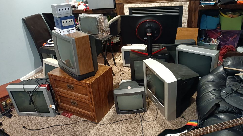 A picture of 10 CRTs scattered around a small area. One CRT has a very large gaming monitor sitting on top of it, and one of them has two gamecubes and a wii stacked on it.