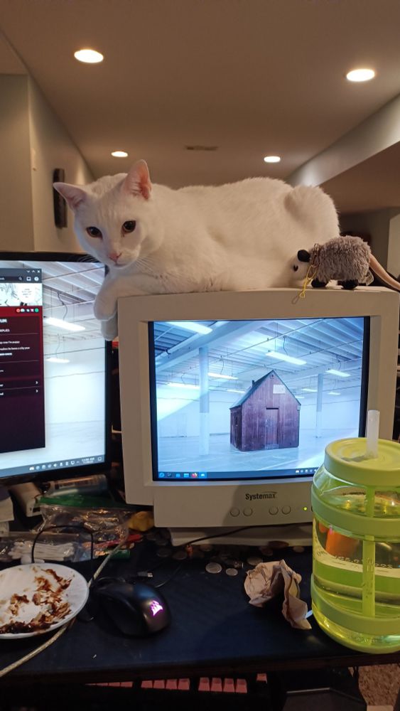 The same white cat perched on top of a CRT computer monitor, looking at the camera, with his arms hanging off. There's also a tiny possum plush