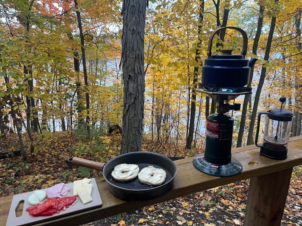 Outside a cabin in the autumn woods. There is a camping burner with a teakettle, French press, a bagel and other food