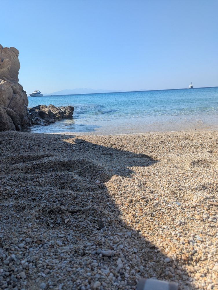 Photograph of a beach full of pebbles, with the water lapping up. The sky is very blue with no clouds