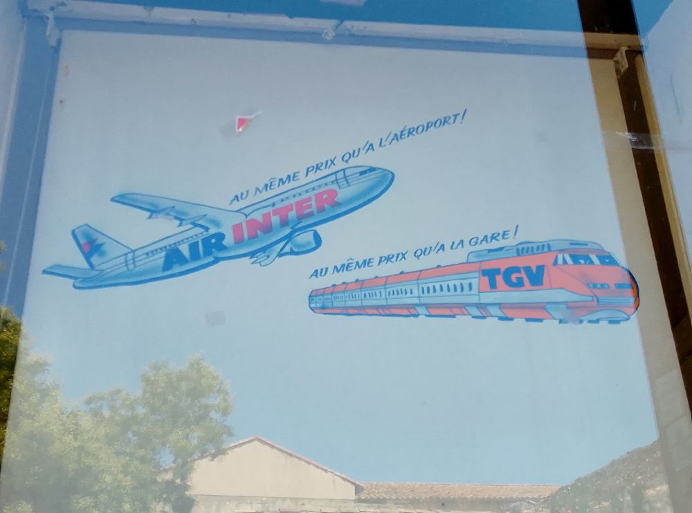 Publicité d'une agence de voyage.
En haut à gauche, un avion siglé Air Inter semble décoller, une inscription se voit au-dessus de l'avion : "Au même prix qu'à l'aéroport !". En dessous à droite, une rame de TGV de couleur orange est surmontée de l'inscription "Au même prix qu'à la gare !".
Dans la fenêtre se reflètent des toitures d'immeubles et quelques arbres.