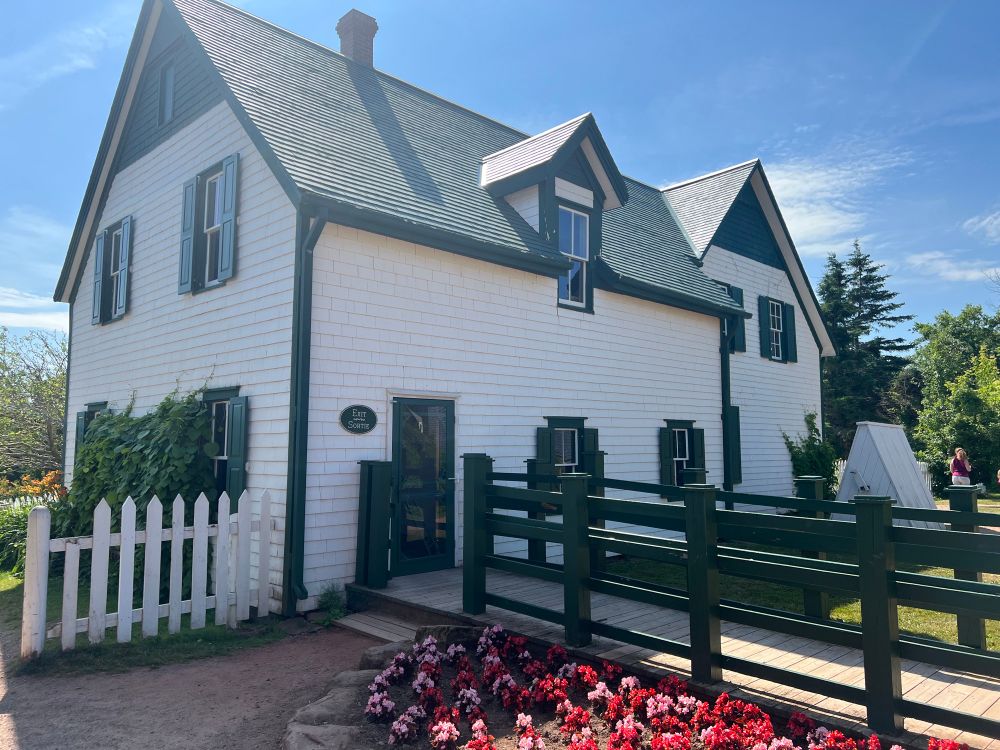 A photo of the back of the Green Gables heritage place house in cavendish, PEI. The house is a large, old-fashioned farmhouse with white siding and green roofing. Overhead the sky is mostly clear with a scattering of clouds; some flowers are growing beside a boardwalk with railings that leads away from the house