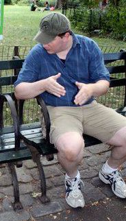 Wrex sitting on a bench in Central Park, NYC. His hands are up and open, close together - as though indicating two things coming together, and his head is turned to his right. He is wearing relaxed fit cargo shorts that are very tight. 