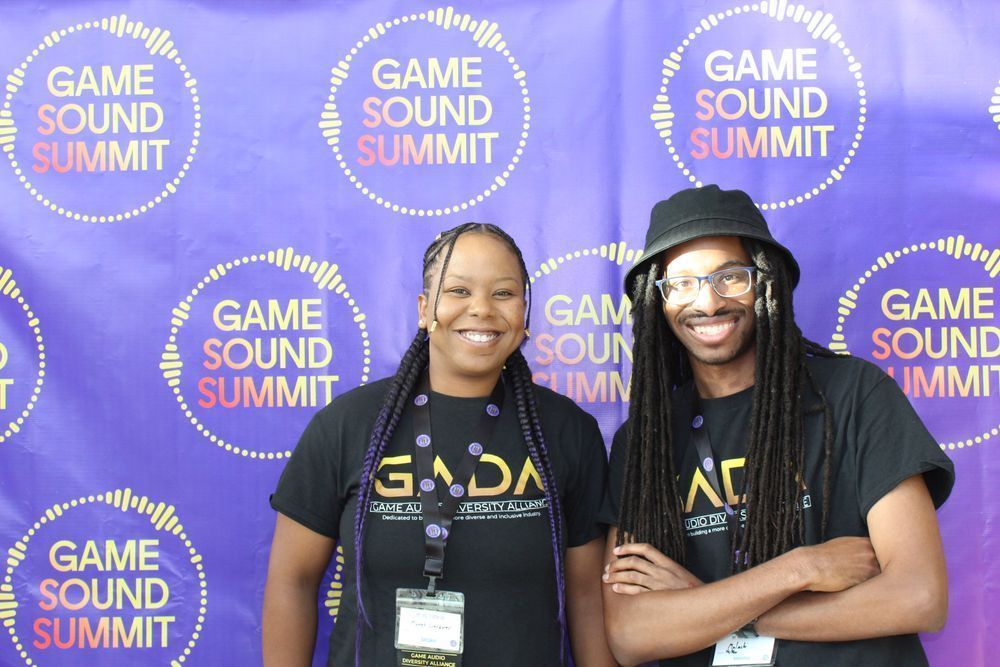 Two people wearing GADA shirts in front of a Game Sound Summit wall.  