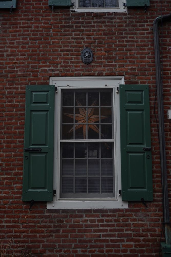 A multi-paned window set in a white frame with green shutters in a red-brick building. Inside the window is a six-pointed star.