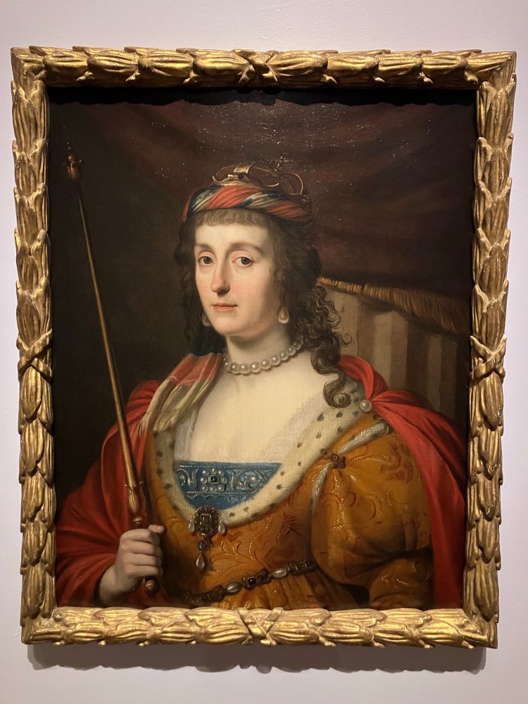 A woman holding a scepter, wearing an orange dress and a red cape.