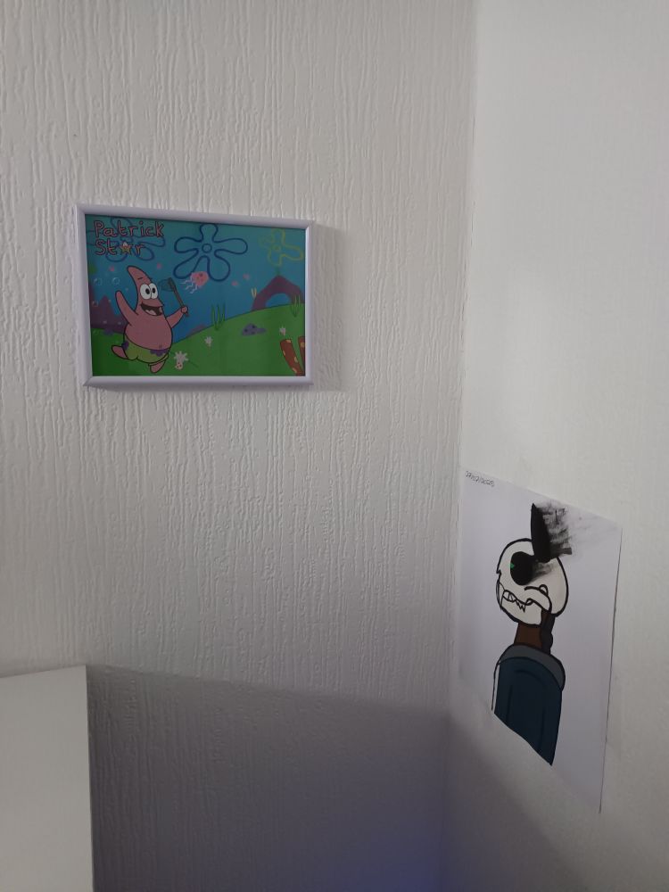 A corner of a white room with 2 pieces of art, on the left is a framed digital illustration of Patrick chasing a jellyfish happily in jellyfish fields from the TV show Spongebob. On the right is an acrylic painting of my sona, a side profile of a cat skull head with smokey eyes and ears, with a wood & metal neck and is wearing a grey and dark blue hoodie. In the bottom left corner there is a white desk edge, and at the bottom a blue light is emanating. 
