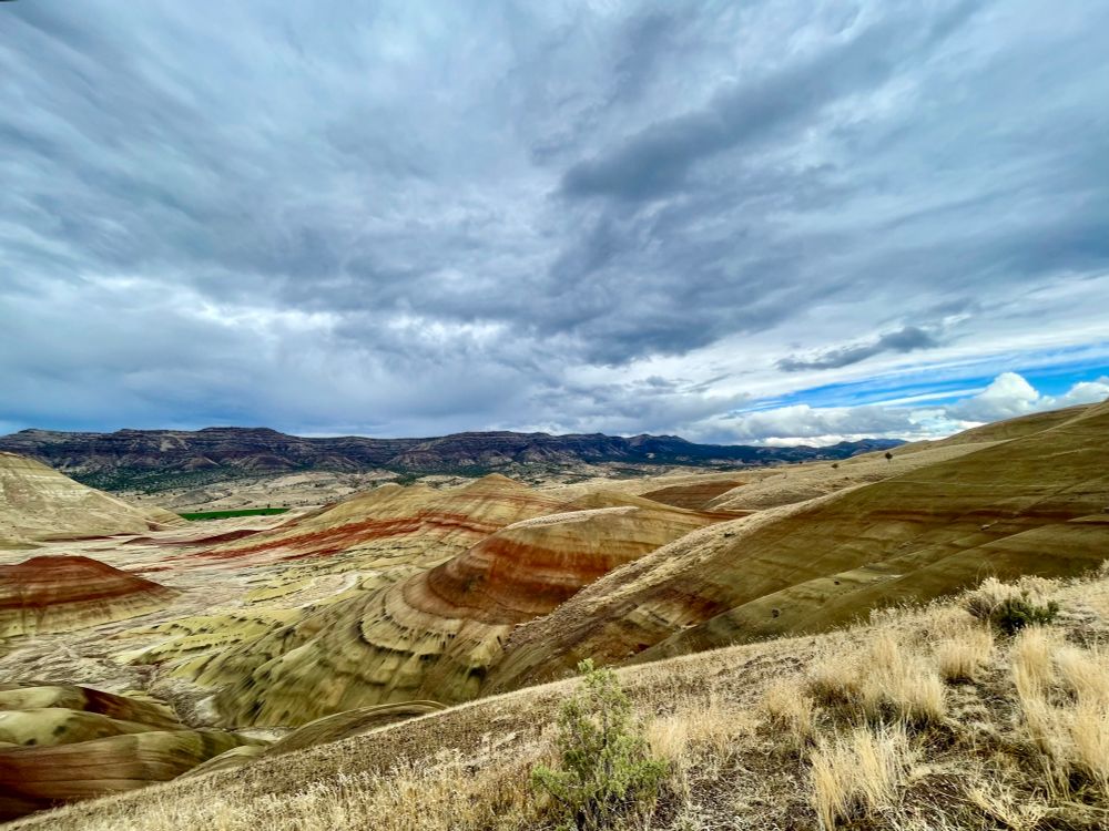 Cloudy sky with small bar of blue sky peaking in background. In foreground there are multiple mounds of the painted hills. These hills have rich green, yellow and orange-red horizontal stripping. Yellow grasses are nearest the camera