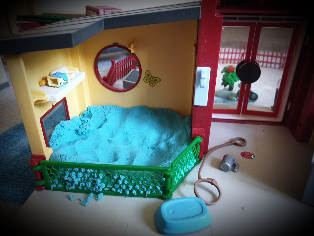 Foto eines Playmobilsets mit Blick in das Innenleben, aktuell dient es als Hundetagesstätte. Zu sehen ist ein Innengehege abgegrenzt mit einem kleinen grünen Zaun, welches mit blauem kinetische Sand gefüllt ist, dieser dient als Wasser. Die Kinder haben den Hunden einen Pool gebaut.
