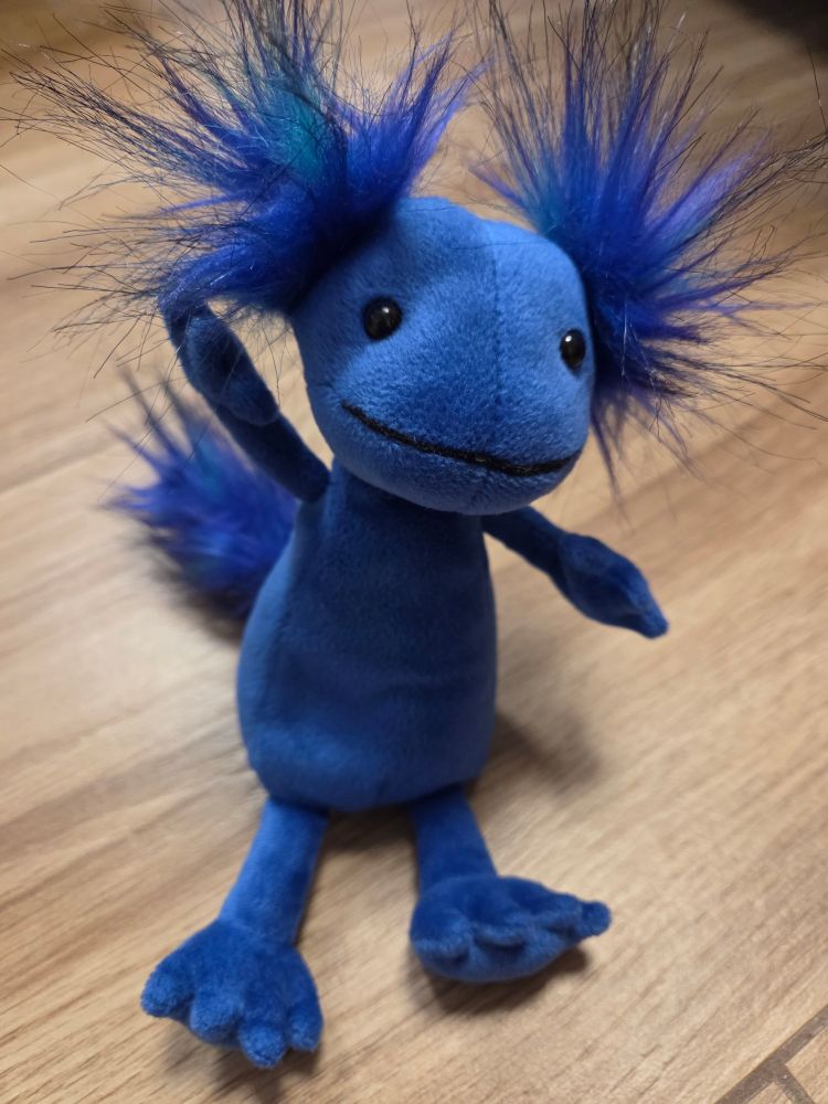 Foto eines Plüschaxolotl.
Das Axolotl ist satt blau, guckt freundlich und winkt mit einem Ärmchen in die Kamera. Besonders niedlich sind zwei Plüschpuschel, die wild rechts und links vom Kopf abstehen und die Kiemen darstellen sollen.