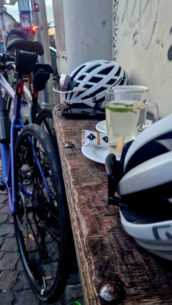 Vor einem Café stehen zwei Räder, auf einem Holzbrett an der Wand stehen ein Kaffee und ein heißer Holunder, daneben liegen Helme