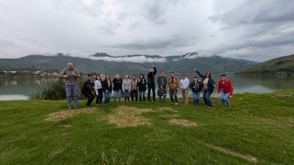 Un grup de persones posant a l'aire lliure davant d'un llac, amb muntanyes verdes i núvols baixos al fons. La majoria dels membres del grup estan fent gestos alegres, com saltar o aixecar els braços. Van vestits amb roba informal i d'excursió, alguns amb jaquetes i altres amb motxilles o càmeres. L'entorn és natural, amb gespa verda en primer pla i el llac reflectint el paisatge. L'ambient sembla fresc i ennuvolat.
