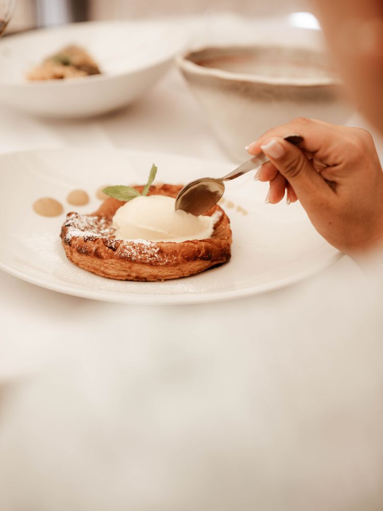 Persona disfrutando de un postre que parece una apetecible tarta de manzada de hojaldre con helado de vainilla, decorado con menta y acompañado de salsas en un plato blanco.
