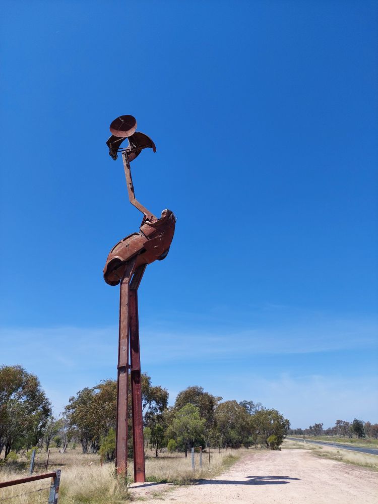 Stanley the big emu, towering over surrounding trees, it's rust red colour contrasts against the sky. Body made from a  VW and the beak from a VW bonnet. 