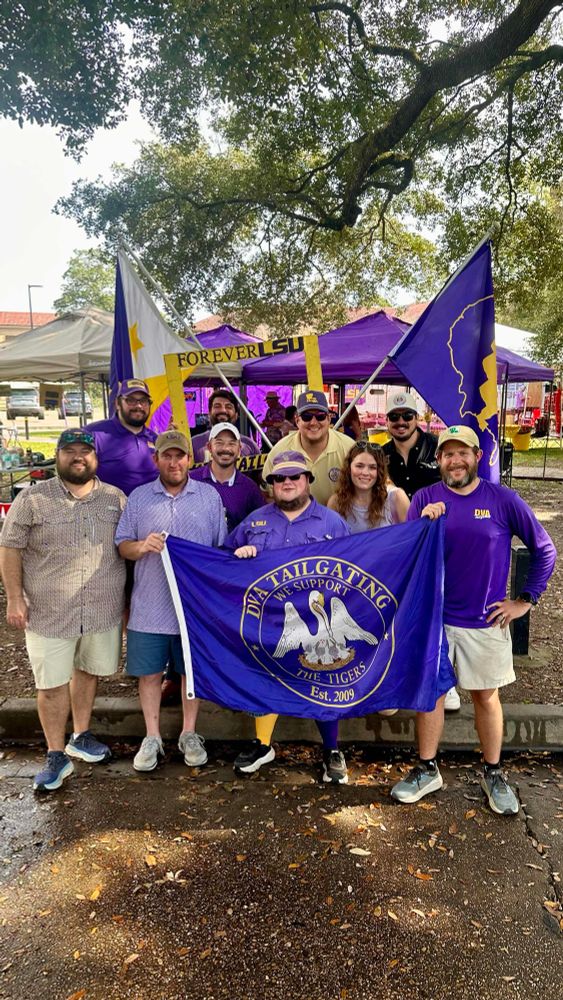 A gaggle of men and a woman posing in from of some flags. In the middle there’s a flag that says “DVA Tailgating - Est. 2009, We Support the Tigers”