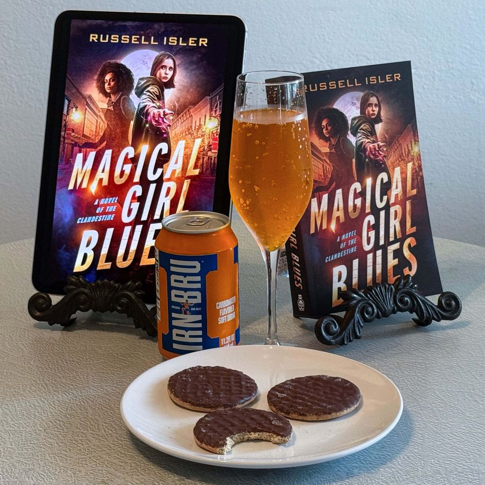 A photo of an ipad displaying the cover of Magical Girl Blues by Russell Isler, a paperback copy of the book and a champaign flute of Irn Bru, a can of the same, paired with McVitie's digestives.