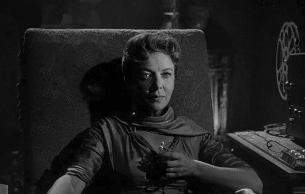 Aging actress in a dark room, rapt with attention, a film projector to the side that's playing her movies (offscreen), which she's watching.