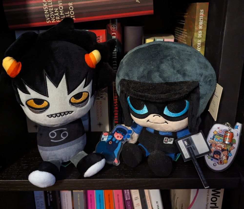 A picture of the same makeship Karkat plushie sitting on my shelf with the makeship Benrey plushie. They both have huge heads. 