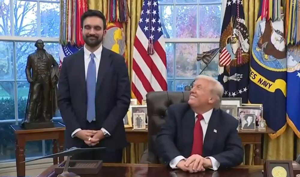 Zohran Mamdani, mayor-elect of New York City, stands beside Donald Trump seated at the Resolute Desk in the Oval Office. The seated man is looking up at the standing man and smiling. The room features large windows, American flags, presidential memorabilia, and framed photographs on the desk.
