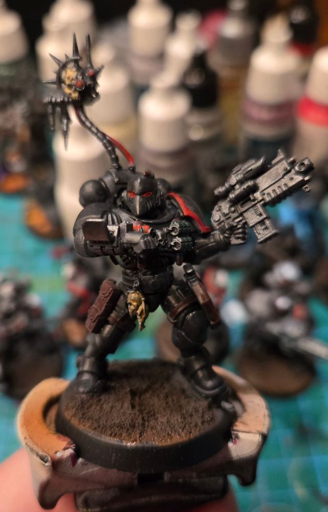 A space marine in stealthy black armor with glowing red lenses and a mechanical skull hovering over his shoulder is looking at a wrist mounted computer and holding a large gun up in his left hand. Front view. 