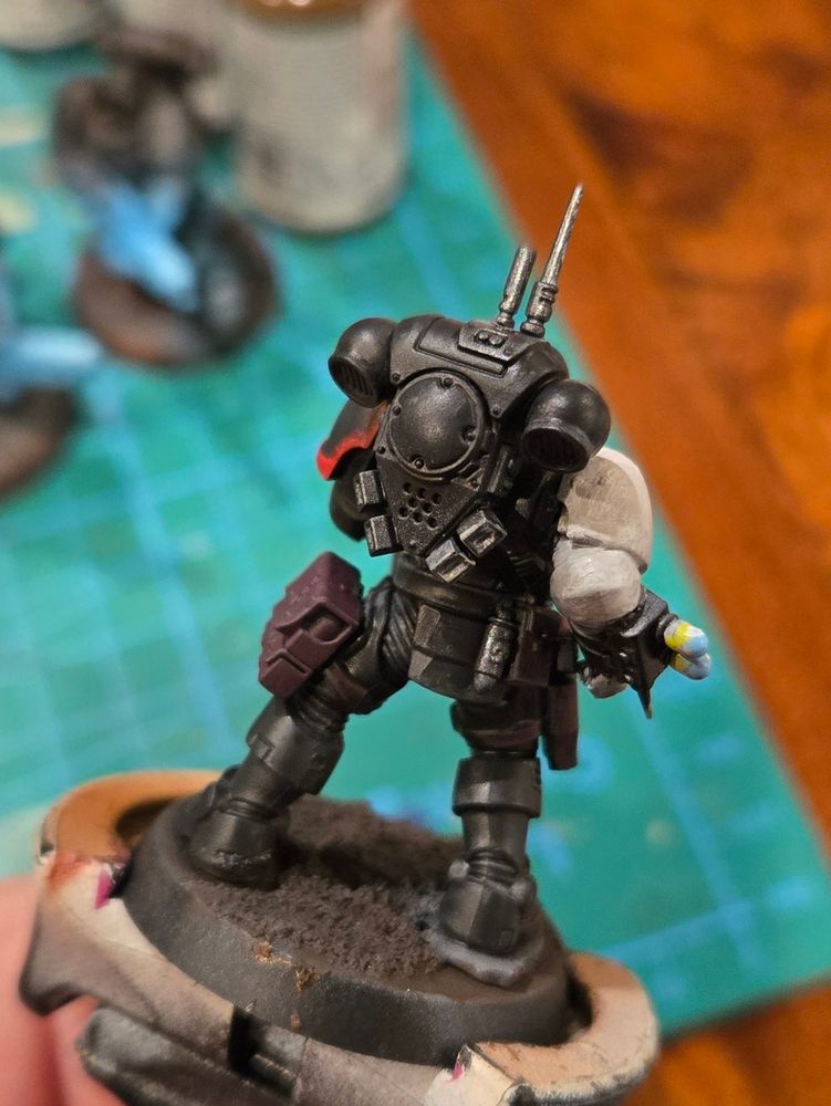 A space marine in stealthy black armor with glowing red lenses. His right arm is white and has a syringe mounted on his right wrist. He is holding a large gun in his left hand and reaching for his holster with his right. Back view. 