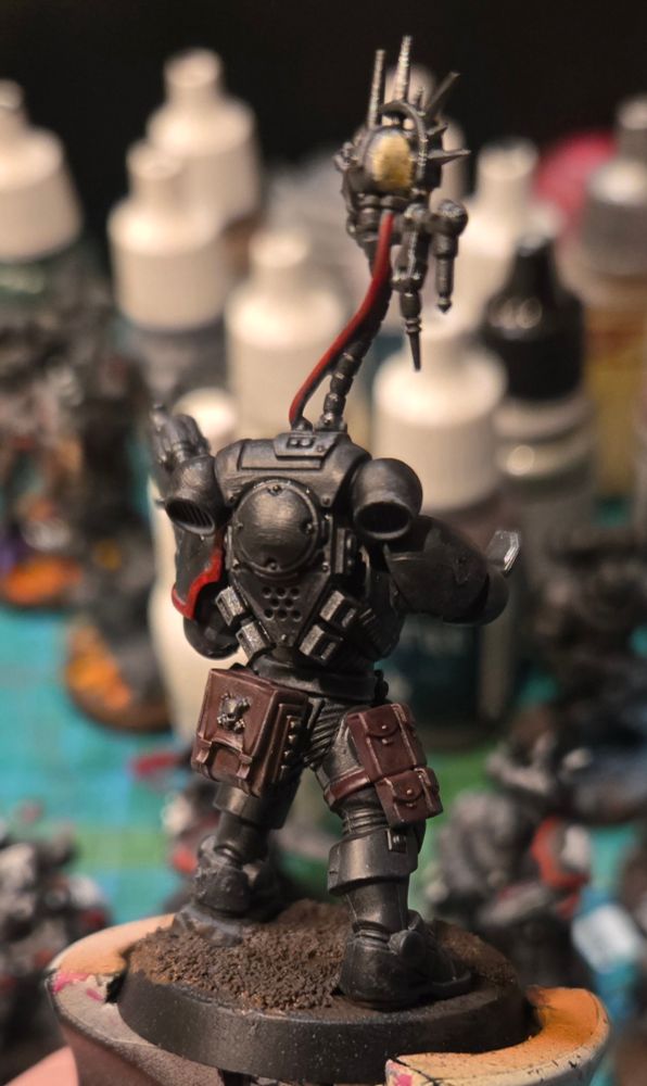 A space marine in stealthy black armor with glowing red lenses and a mechanical skull hovering over his shoulder is looking at a wrist mounted computer and holding a large gun up in his left hand. Back view. 