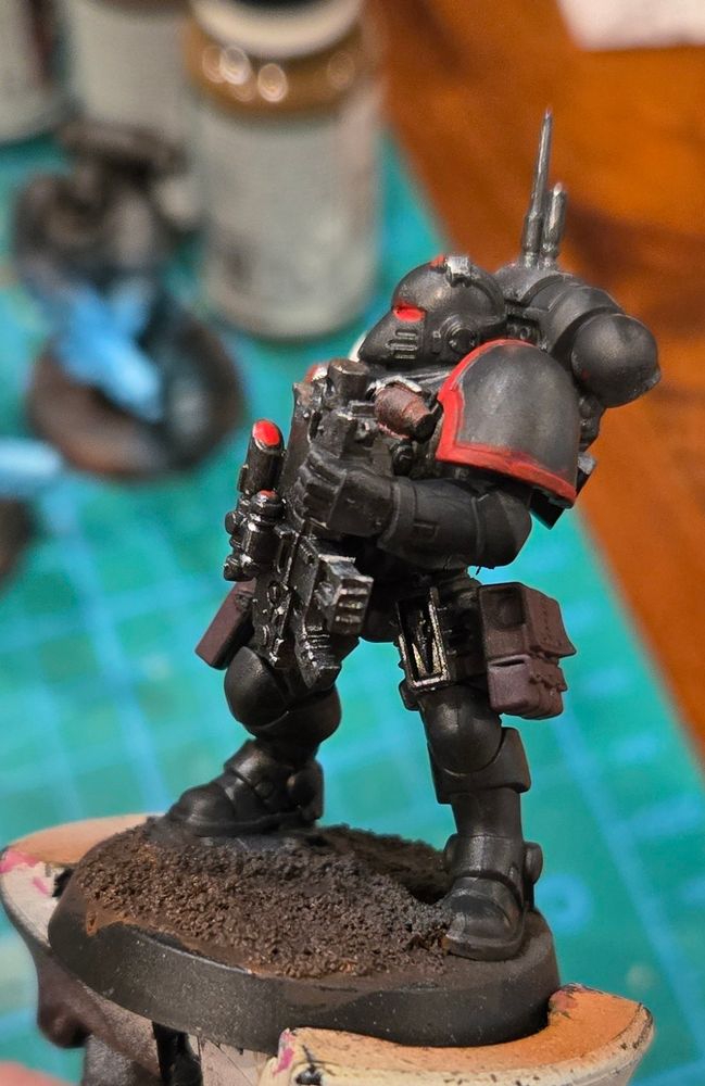 A space marine in stealthy black armor with glowing red lenses. His right arm is white and has a syringe mounted on his right wrist. He is holding a large gun in his left hand and reaching for his holster with his right. Side view. 