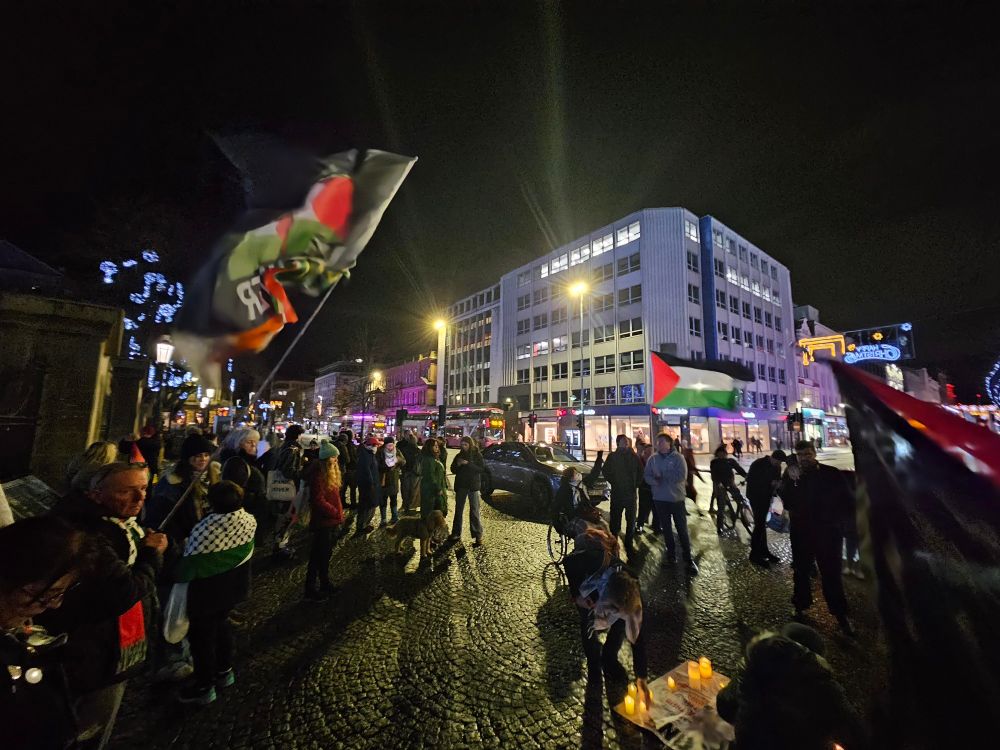 People gathered on the cobbles in front of Belfast City Hall, some waving Palestine flags. A banner with lit candles rests on the ground.