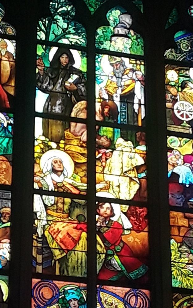 A detail from a window in Prague's St. Vitus cathedral, painted by Alphonse Mucha, showing the spread of Christianity among the Bohemian Slavs (Czechs). To the top are the two Apostles to the Slavs, Saints Cyril and Methodius, baptizing Borivoj. In the lower half Saint Ludmila prays with her grandson, Saint Wenceslas. Cyril wears a monk's habit in somber brown, while Methodius is garbed in a bishop's vestments. Ludmila arms outspread in prayer, is wearing a gown of light yellow-green, belted with gold, and a red apron, while young Wenceslas wears a vibrant red tunic and has his hands clasped in front of him.