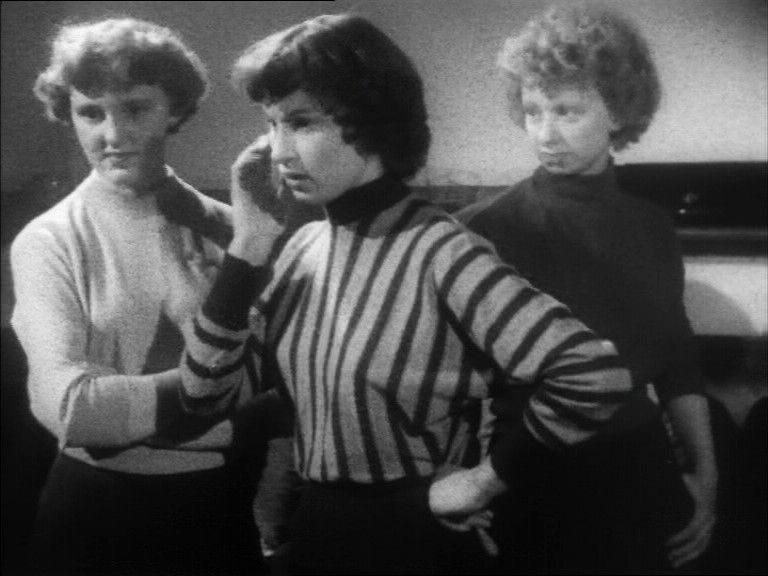 Still image. Three young British women in sweaters look intense.