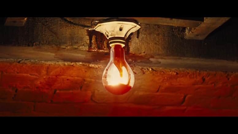 Still image. An illuminated light bulb with blood on it in front of a red brick wall.