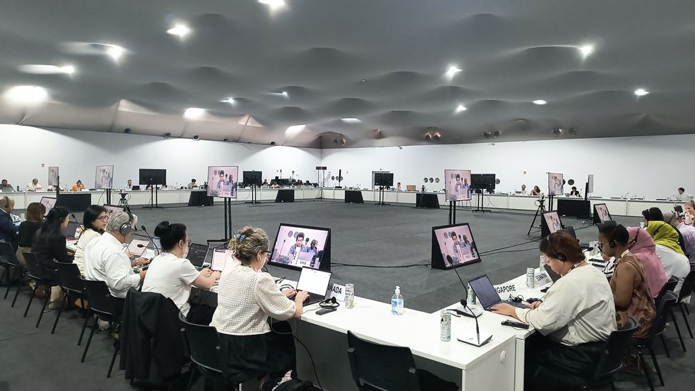 Uma sala ampla, de paredes brancas, com mesas colocadas lado a lado formando a borda de um quadrado. Aproximadamente 25 lugares em cada lado. Está parcialmente ocupada. No interior deste quadrado algumas telas de televisão ligadas. 