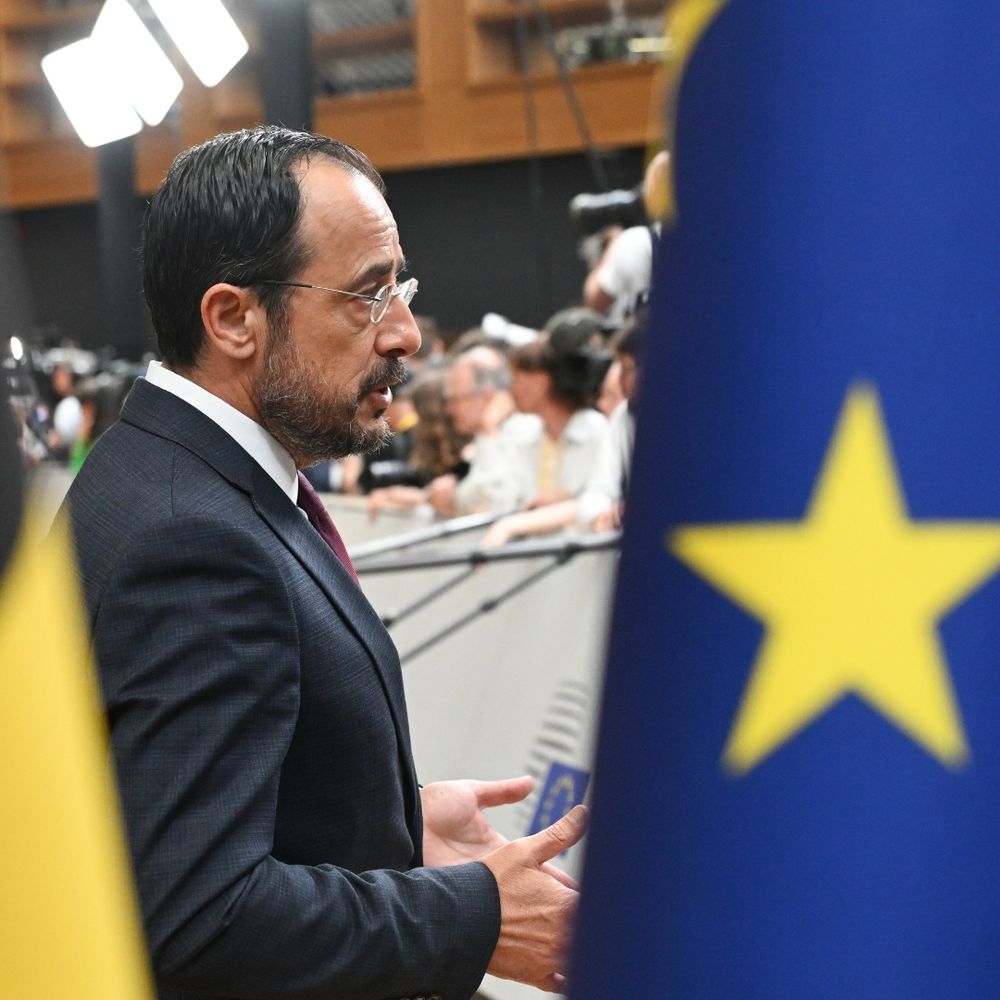 At the front part of what seems to be the European flag. You can see a yellow star on a blue background. In the back a man in a grey suit talking.